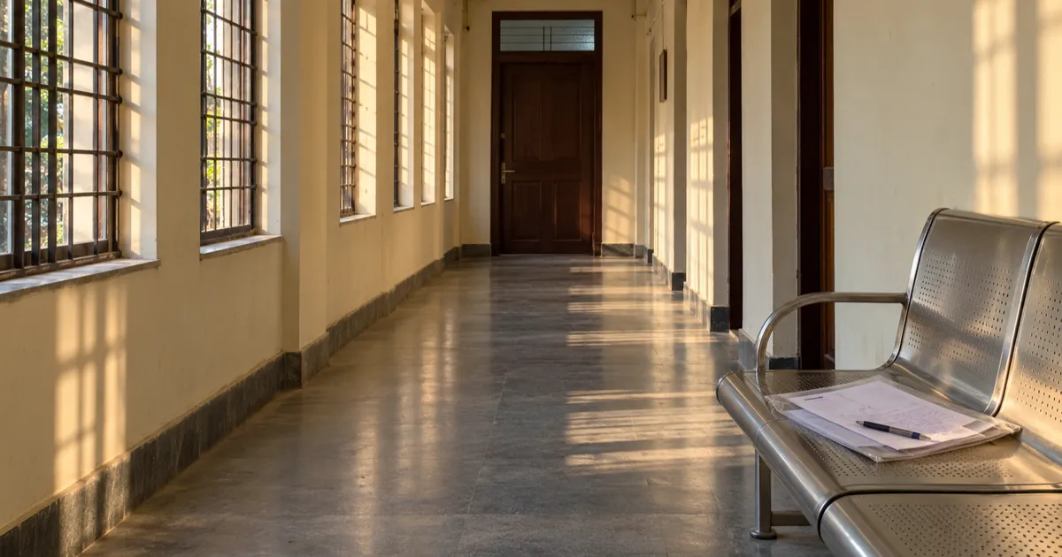A clear plastic folder holding a handwritten complaint and photocopied receipts rests on a steel bench outside a closed wooden office door in a long cream-walled college hostel corridor.