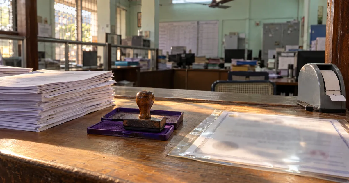A wooden service counter in a municipal citizen service centre holds a stack of stapled forms, a rubber stamp on an ink pad and a certificate in a clear plastic sleeve, with a queue token machine and a green wall behind under a ceiling fan.