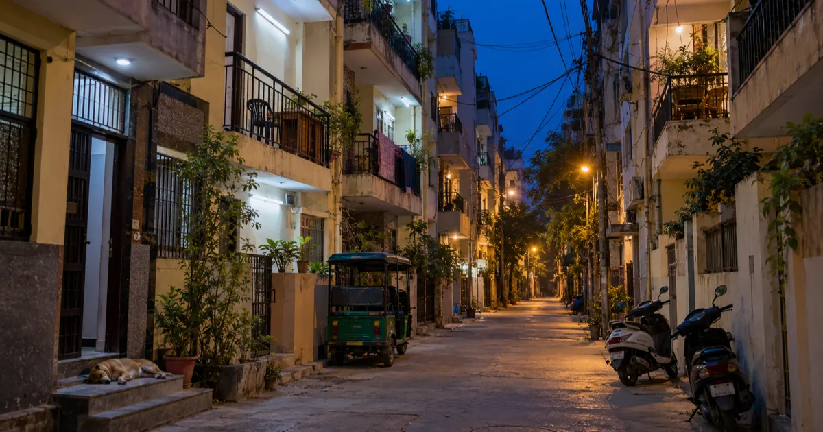 A narrow north Delhi lane at dusk lined with four- and five-storey residential buildings, small balconies with study desks and lit tube lights, tangled overhead cables and a parked green e-rickshaw below.