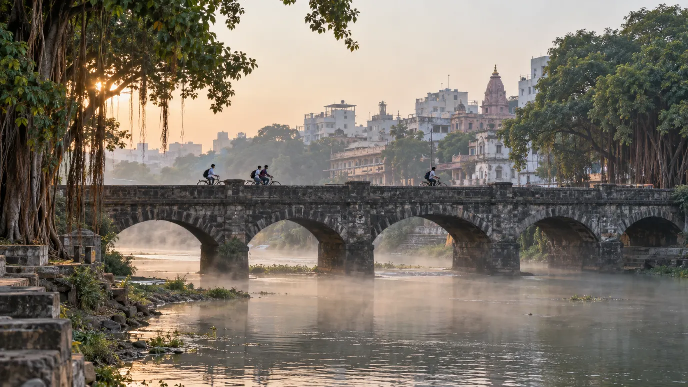 Cyclists cross an old stone arch bridge over a misty river at sunrise, with banyan trees and old city buildings behind in Pune