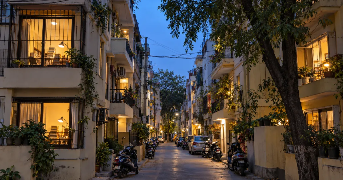 A narrow residential lane in Old Rajinder Nagar at dusk, lined with four-storey buildings whose small balconies hold study tables and desk lamps, with parked scooters and loose overhead cables.
