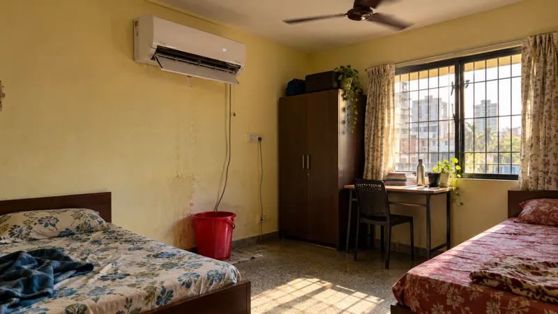 A broken white split air conditioner with its front panel hanging open drips into a red bucket in a twin-sharing PG room with two single beds and a ceiling fan, lit by harsh afternoon sun.