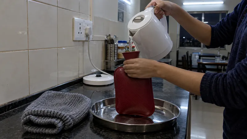 Student pouring water from a white electric kettle into a red rubber hot water bag held over a steel tray on a hostel pantry counter, a grey knitted cover beside it