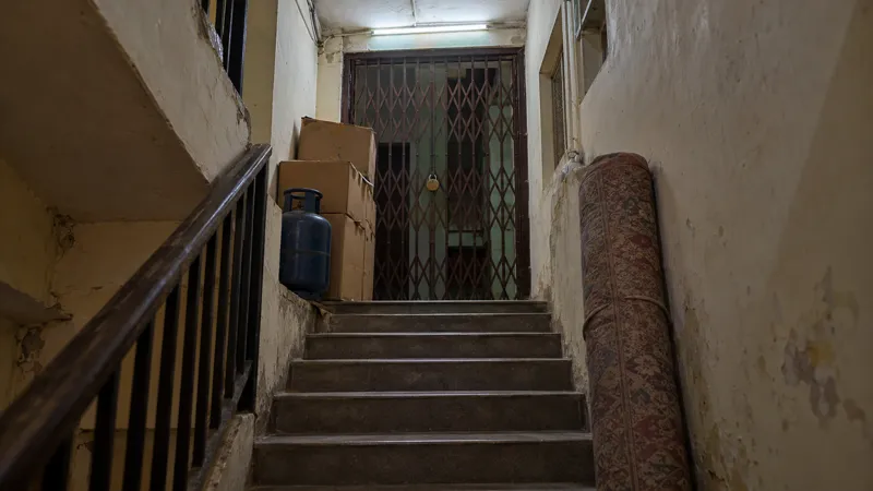 Cardboard cartons, a blue gas cylinder and a rolled mattress block the narrow staircase landing of a converted PG house, below an iron grille gate shut with a brass padlock under a tube light.