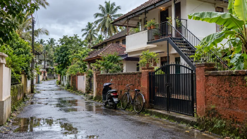 A narrow wet residential lane in a Kerala suburb with laterite compound walls, tiled-roof two-storey houses, an outside steel staircase to an upper room, and a bicycle and scooter parked by an iron gate under an overcast sky.