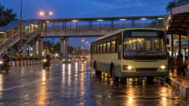 A plain town bus pulling into a roadside shelter on a wide Coimbatore road at dusk, with a pedestrian foot overbridge behind it, passing two-wheelers and street lights reflecting on wet asphalt.