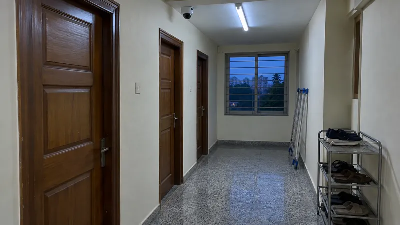A white dome CCTV camera on the ceiling of a narrow PG corridor at dusk, above closed wooden doors, a shoe rack with sandals and a drying stand by the window.