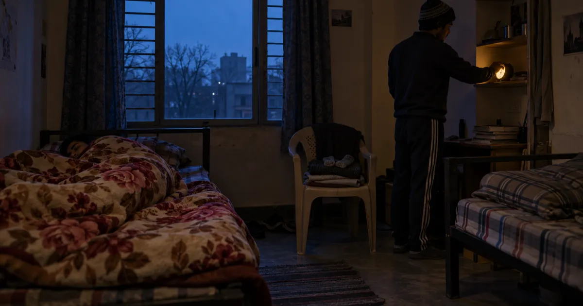 In a dark hostel room before dawn, a student in a woollen cap and track pants switches off a glowing alarm clock on a shelf while his roommate sleeps under a floral quilt, with folded warm clothes on a plastic chair.